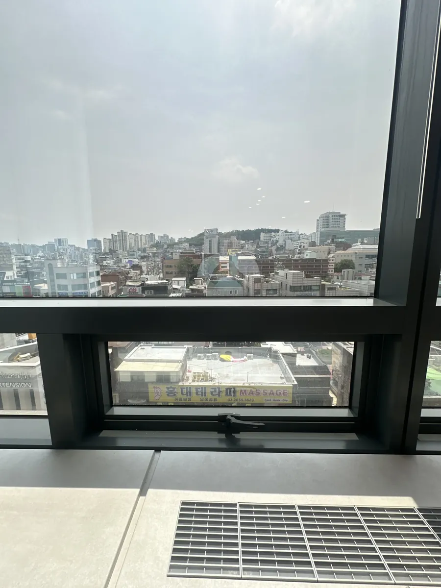 고층 사무공간 환기창 경첩 조정 중 도심 상가 간판이 보이는 창밖 전경